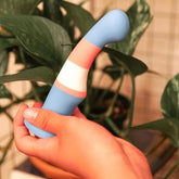 A hand holds the Avant Blush Avant P2 Silicone Pride True Blue Dildo, made from body-safe silicone and featuring blue, pink, and white stripes; leafy green plants appear blurred in the background.